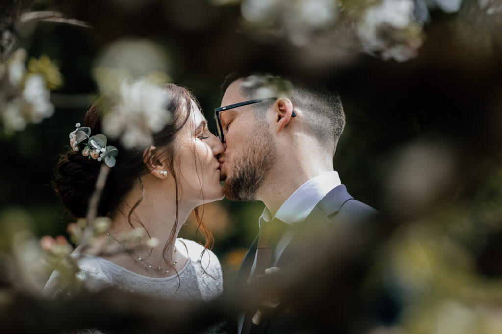 Brautpaar küsst sich und die beiden Gesichter werden sehr schön im Foto eingerahmt von Ästen und Blüten eines Apfelbaums