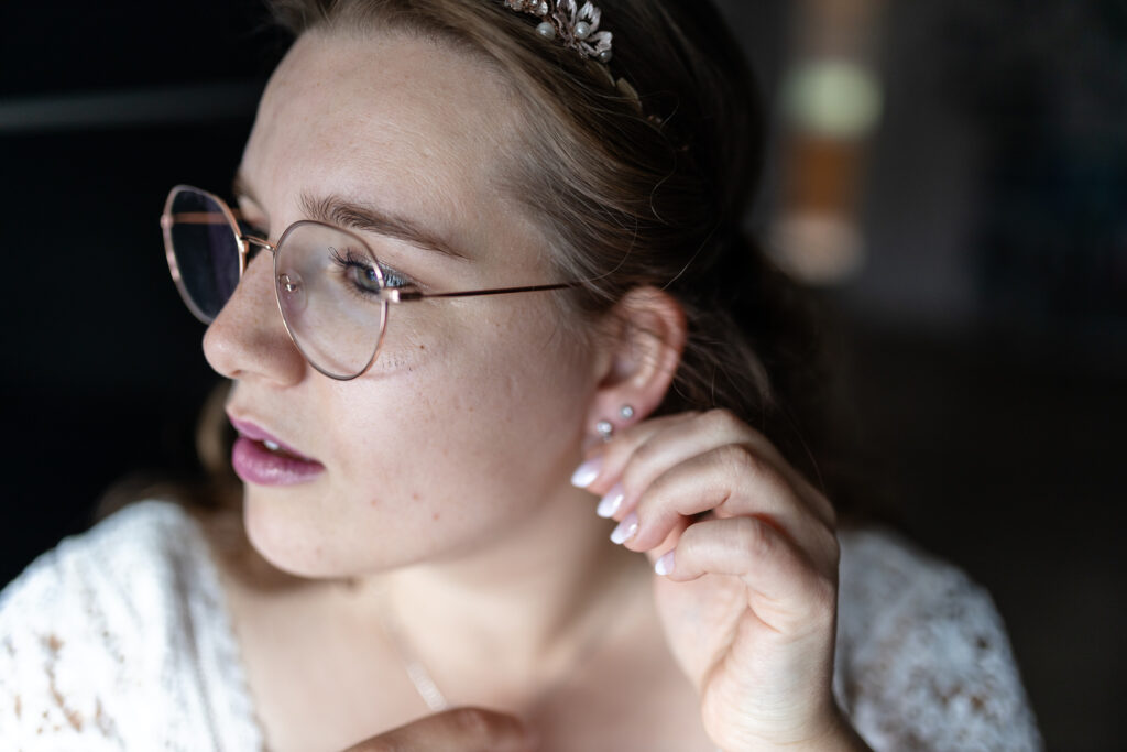 Getting Ready Portrait Kopf Braut: Die Braut steht im wunderschönen Fensterlicht und schaut nach ihren Ohrringen