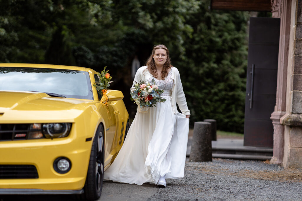 Die Braut geht vor der Kirche an dem gelben Ford Mustang, dem Hochzeits-Auto, vorbei.