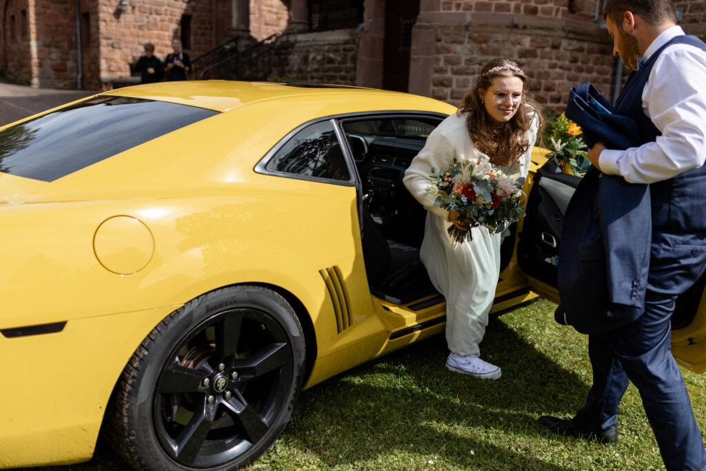 Der Bräutigam hält der Braut die Autotür des gelben Ford Mustang (Hochzeits-Auto) auf und die Braut steigt im Moment aus.