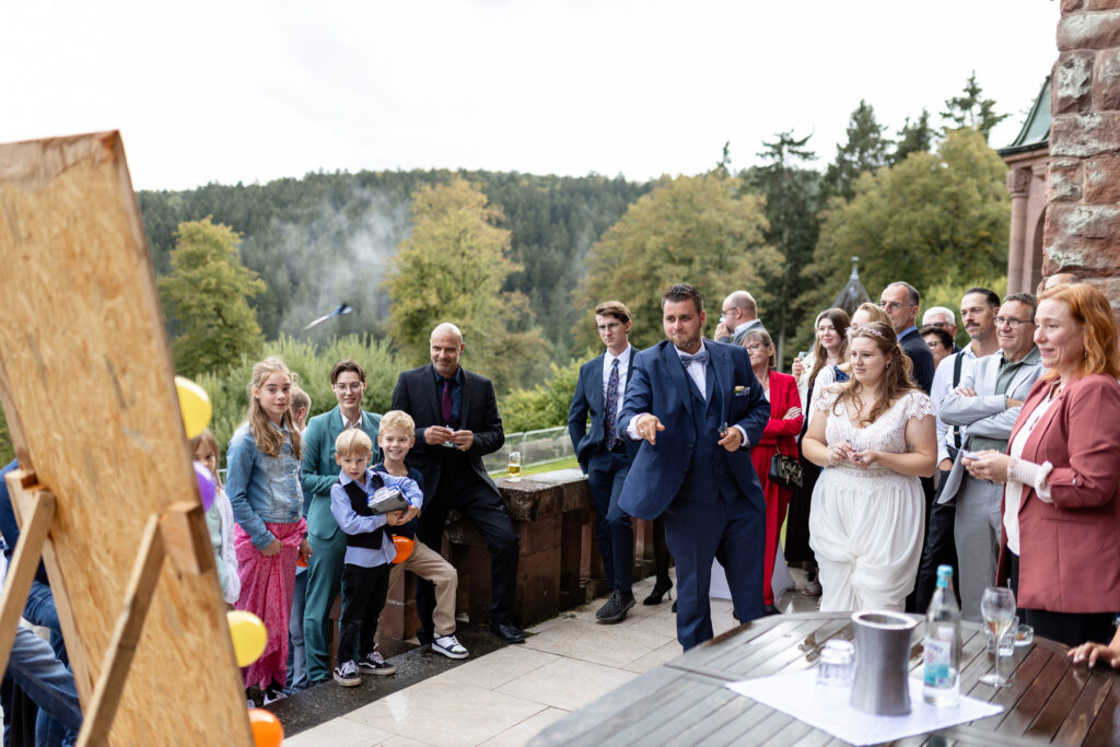 Der Bräutigam wirft einen Dart auf die Zielscheibe des Geschenke-Dart-Spiels seiner Hochzeit. Die Gäste sind drumherum versammelt und schauen dabei zu.