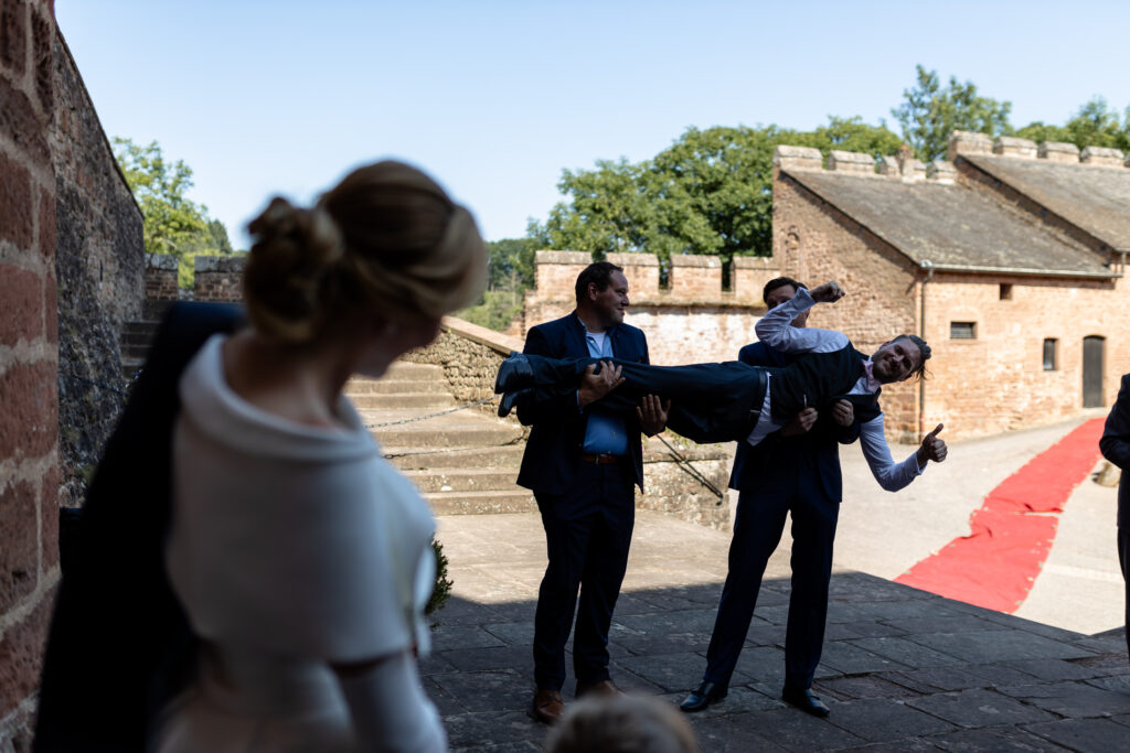 Der Bräutigam wird von 2 Mann hochgehoben, während die Braut zusieht