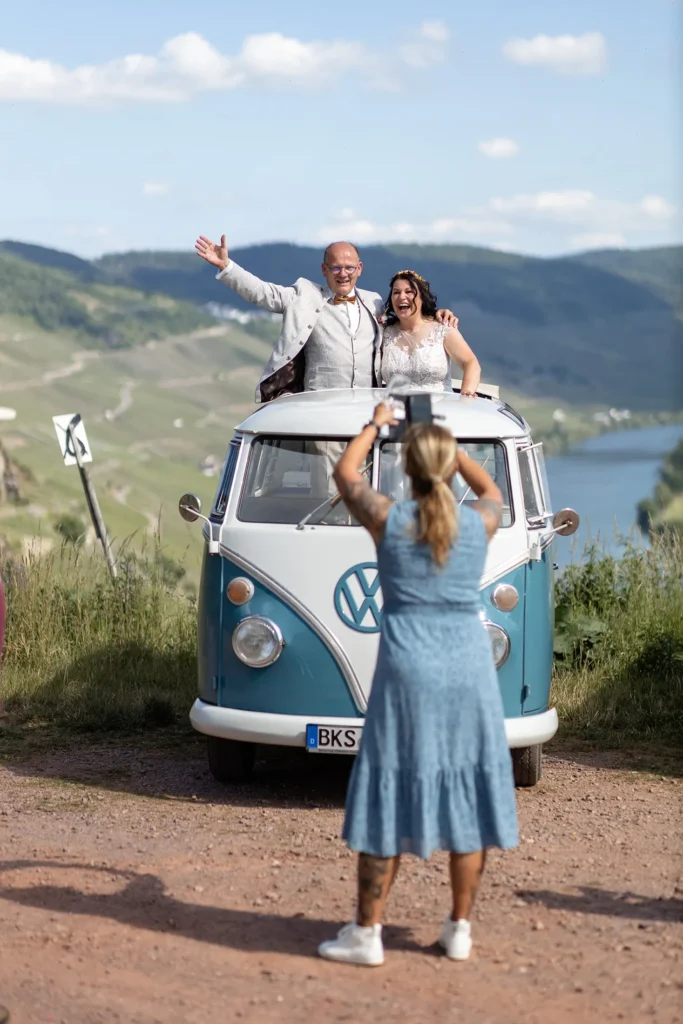 Hochzeitsfotograf Bernkastel Trier - Im Vordergrund unscharf: Ein Gast, der ein Handy Foto vom Brautpaar macht, das gerade aus dem offenen Dach eines T1 Bullis herausragt. Der Bus und das Brautpaar sind scharf.