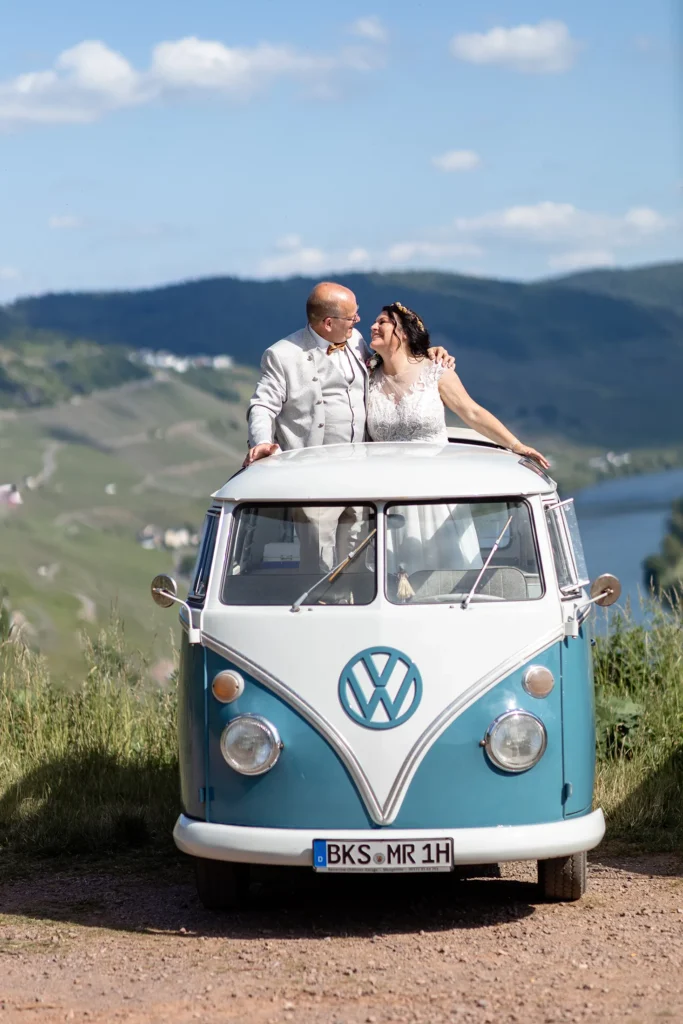 Hochzeitsfotograf Bernkastel Trier - Vordergrund: Brautpaar umarmt sich und ragt aus dem offenen Dach des T1 Bullis heraus. Im Hintergrund sieht man die Mosel und das Tal bei Bernkastel in der Unschärfe.