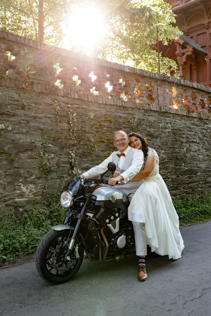 Hochzeitsfotograf Bernkastel Trier - Brautpaar-Shooting vor einer Mauer mit schöner Lichtstimmung. Der Bräutigam sitzt auf seinem Motorrad und die Braut sitzt hinter ihm und hält sich an ihm fest.