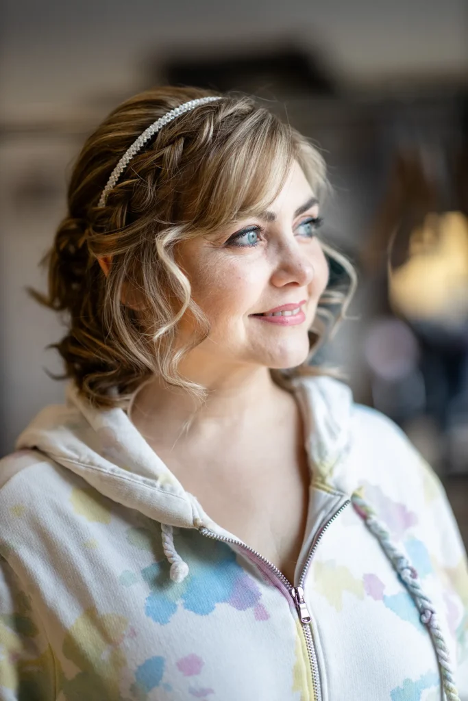 Hochzeitsfotograf Luxemburg - Portrait in schönem Licht: Die Braut steht mit fertiger Frisur am Fenster des Friseursalons und lächelt.