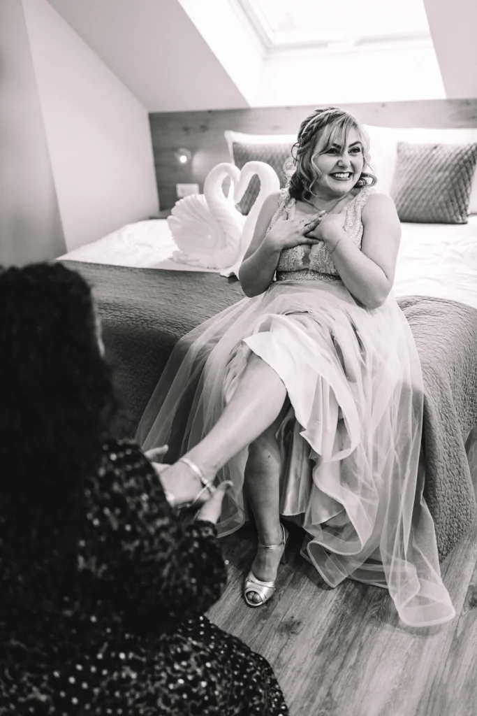 Hochzeitsfotograf Luxemburg - Schwarz-weiss Aufnahme der Braut, wie sie lachend auf dem Bett sitzt, während ihre Freundin ihr mit den Schuhen hilft.