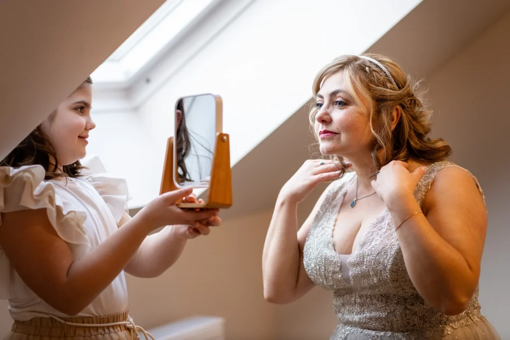 Hochzeitsfotograf Luxemburg - Die Tochter hält der Braut am Fenster einen kleinen Spiegel vors Gesicht und die Braut richtet noch einmal schnell die Haare, bevor es zum First Look geht.