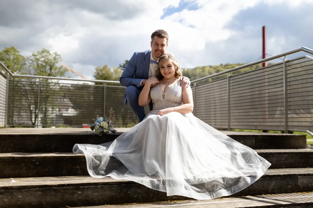Hochzeitsfotograf Luxemburg - Brautpaar Foto: Brautpaar sitzt im Park in Diekirch auf einer Treppe und posiert. Im Hintergrund erkennbar: Der Rote Träger der Brücke über die beide jeden Tag zur Arbeit fahren.