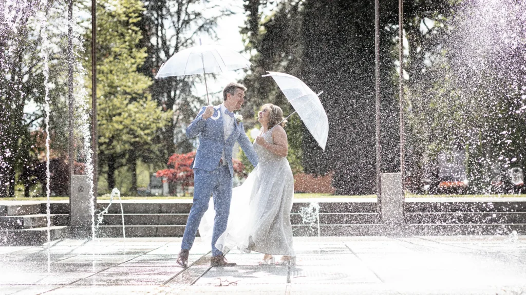 Hochzeitsfotograf Luxemburg - Das Brautpaar hat transparente Regenschirme und posiert in einem Brunnen, in dem das Wasser meterhoch spritzt.
