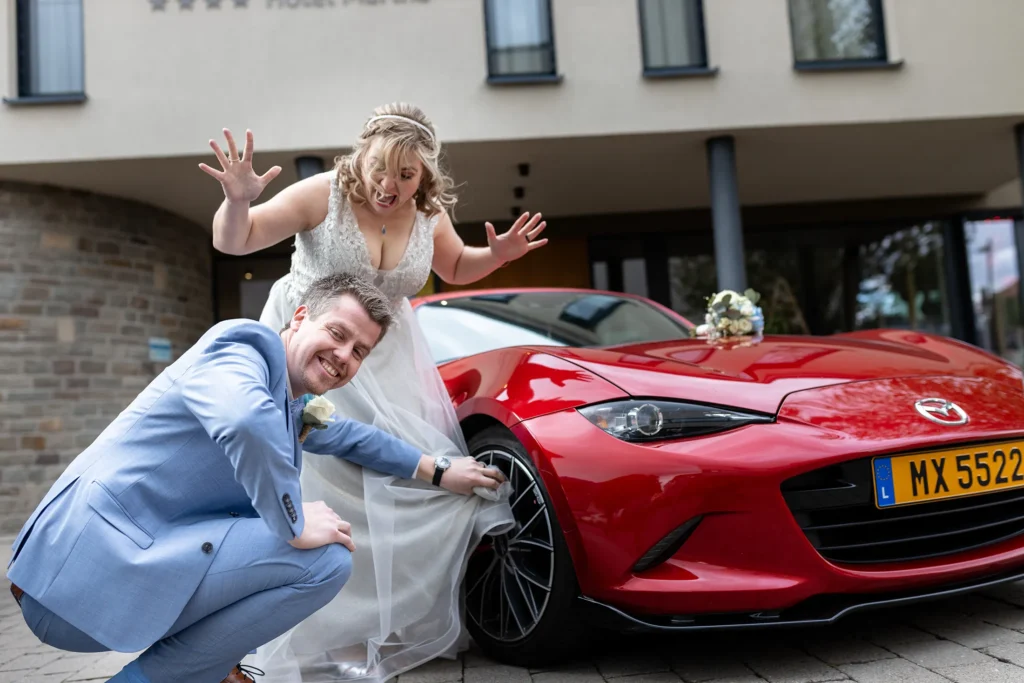 Hochzeitsfotograf Luxemburg - Der Bräutigam wischt mit dem Brautkleid seiner Frau die Felgen seines Sportwagens und grinst in die Kamera, während die Braut sich darüber aufregt und die Arme vor Schreck hochreisst.
