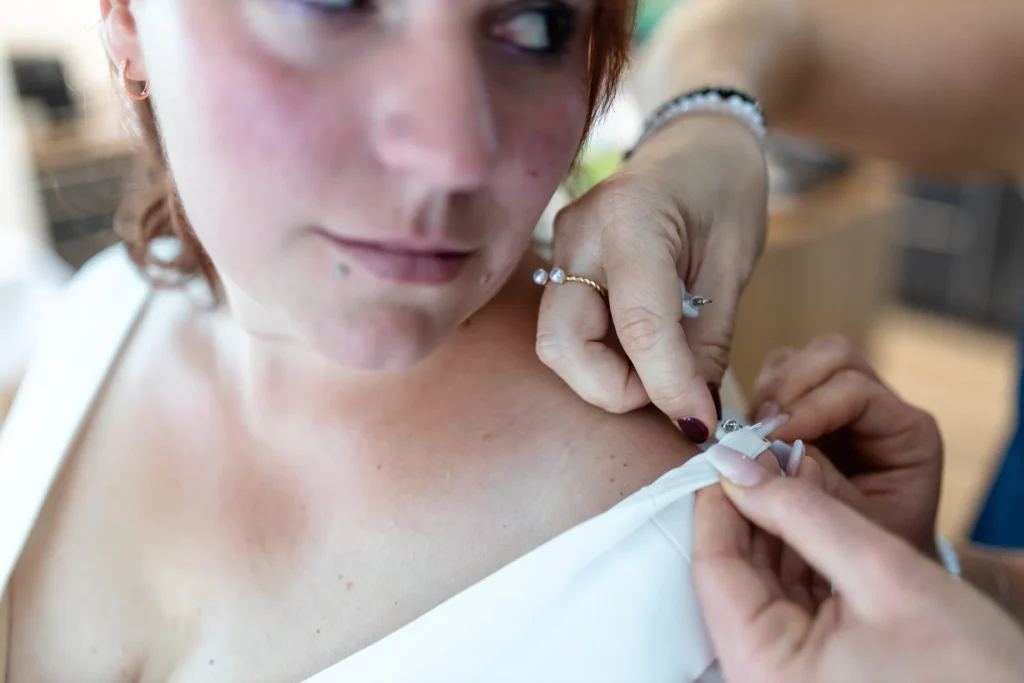 Hochzeitsfotograf Merzig Trier - Interessante Perspektive: Man sieht den angeschnittenen Kopf der Braut und ihre Schulter. Eine Brautjungfer näht schnell noch etwas am Halter des Hochzeitskleids