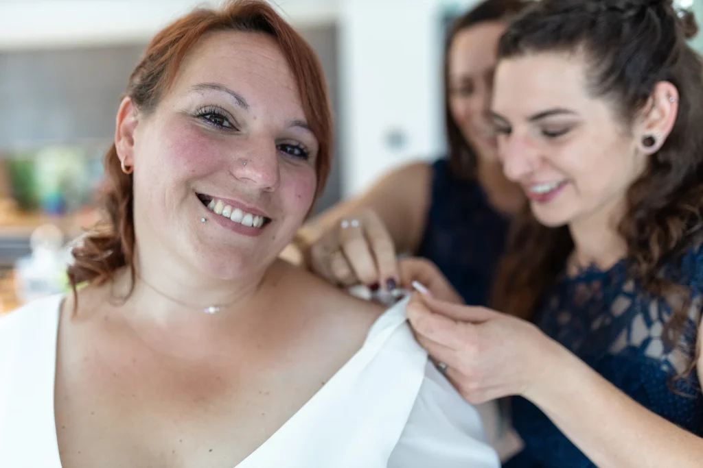 Hochzeitsfotograf Merzig Trier - Braut im Vordergrund. Im Hintergrund sieht man unscharf, dass zwei Brautjungfern noch etwas am Halter des Hochzeitskleis ausrichten.