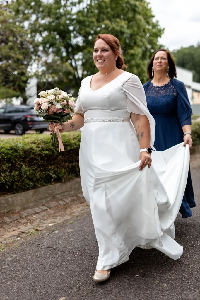 Hochzeitsfotograf Merzig Trier - Die Braut geht schnellen Schrittes auf dem Weg zum Eingang der Kirche. Eine Brautjungfer hält hinter ihr das Brautkleid hoch.