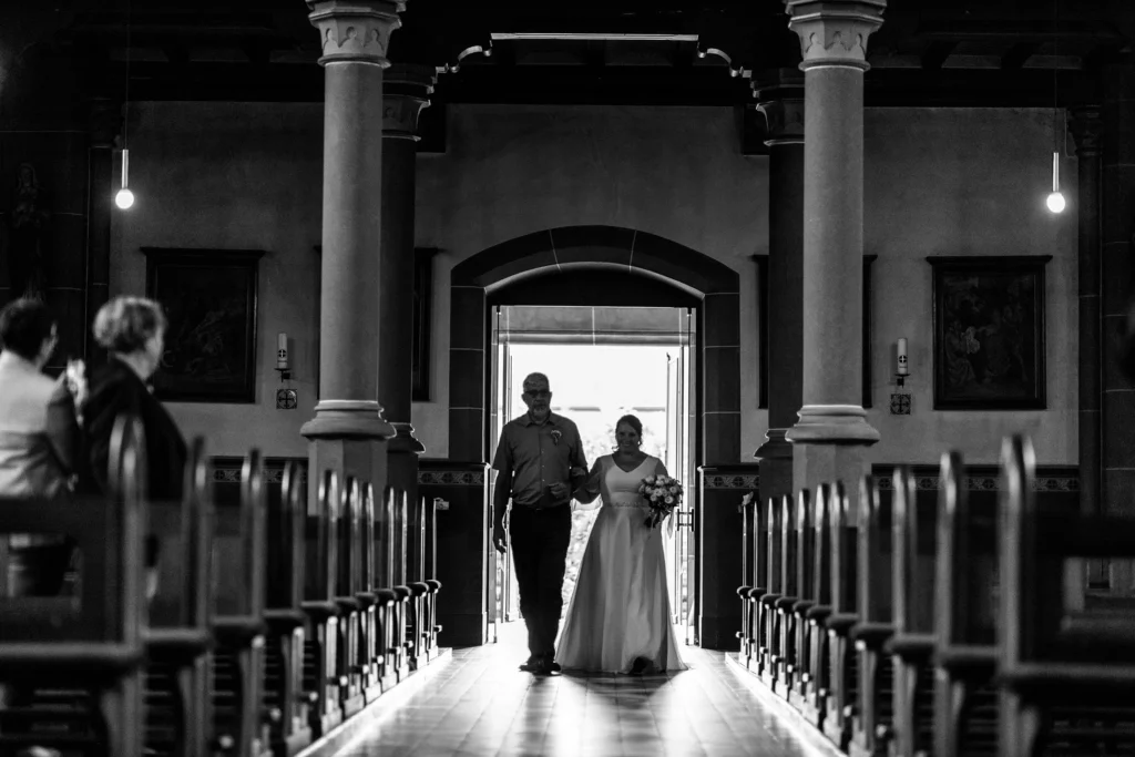 Hochzeitsfotograf Merzig Trier - Eindrucksvolles schwarz-weiss Bild, wie die Braut von ihrem Vater gerade durch die Tür in die Kirche kommt und durch den Mittelgang geführt wird.