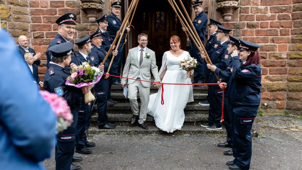 Hochzeitsfotograf Merzig Trier - Das Brautpaar direkt auf den Treppen des Kircheneingangs, während die Feuerwehr Spalier steht.,