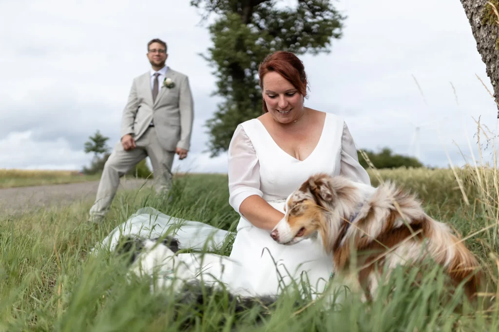 Hochzeitsfotograf Merzig Trier - Die Braut im Vordergrund streichelt ihren Hund, während der Bräutigam aus dem Hintergrund zuschaut.