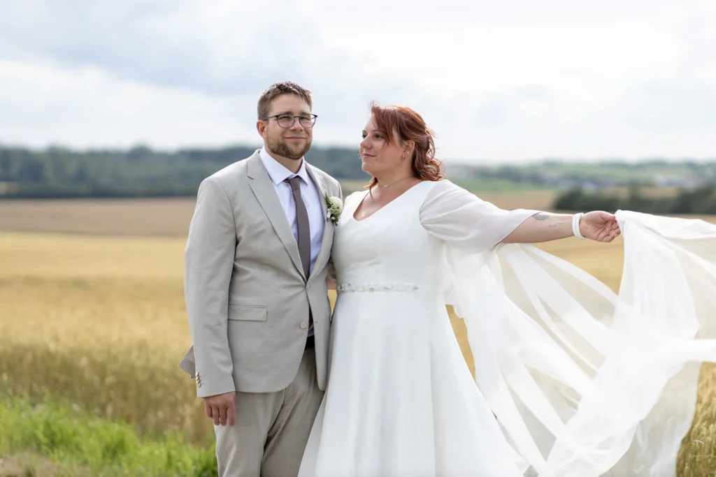 Hochzeitsfotograf Merzig Trier - Die Braut streckt die Arme aus und zeigt ihren langen Schleier beim Brautpaar- Shooting auf einem Feld.
