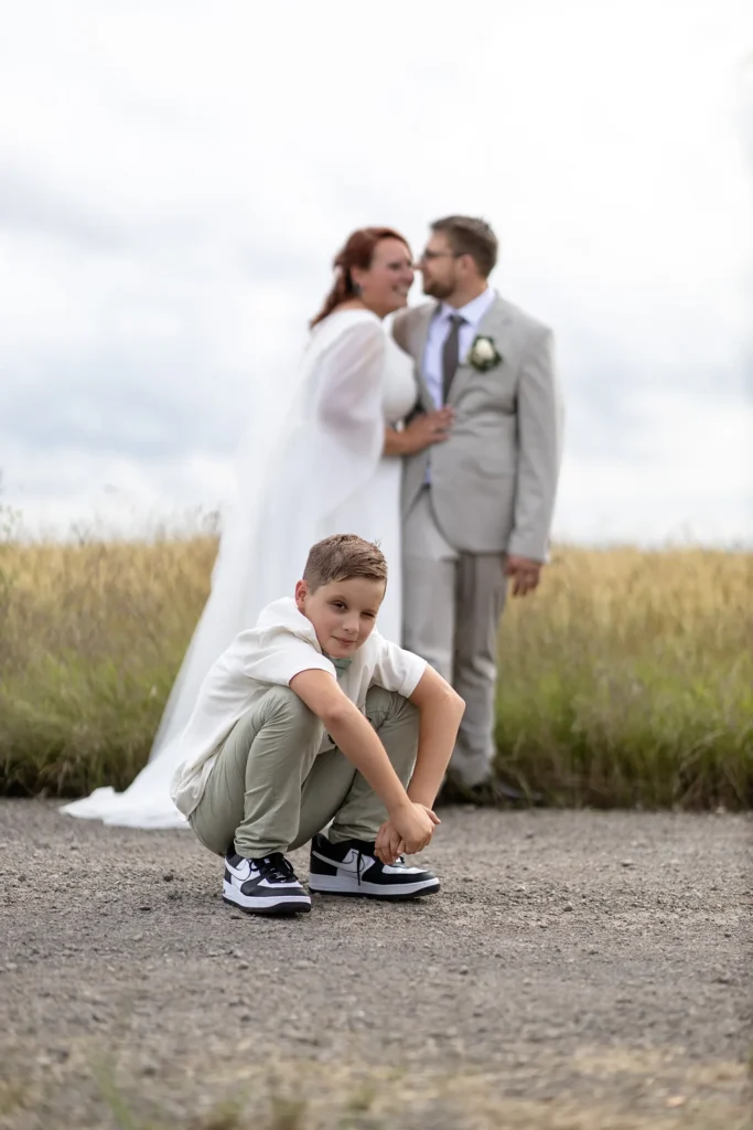 Hochzeitsfotograf Merzig Trier - Portrait des Sohns vom Brautpaar auf offenem Feld in Merzig. Das Brautpaar steht im Hintergrund und lächelt.