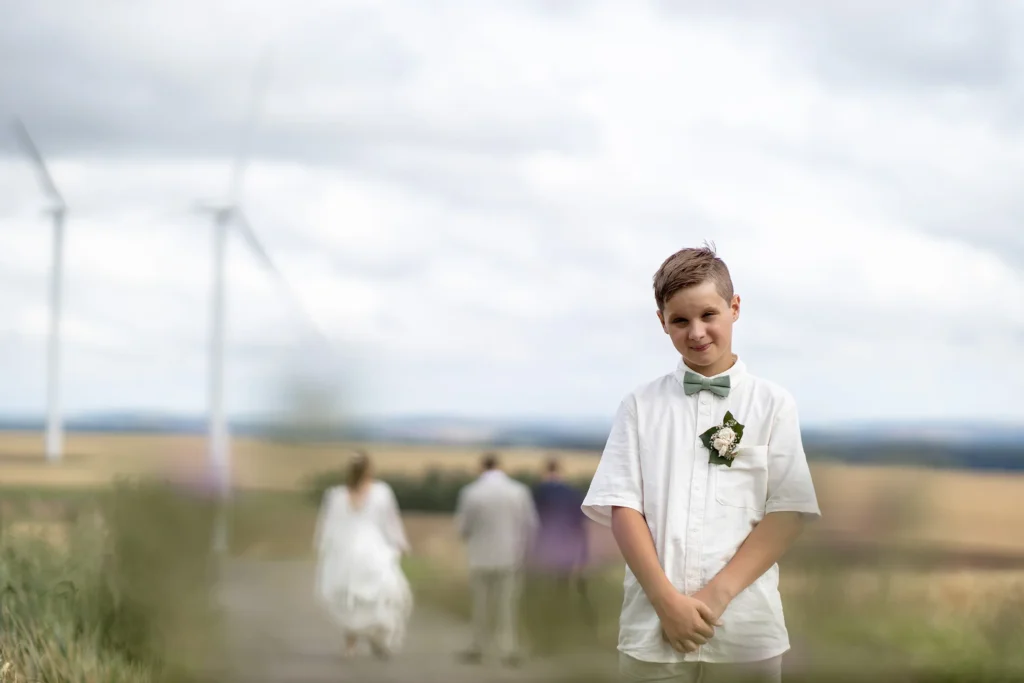 Hochzeitsfotograf Merzig Trier - Der Sohn des Brautpaars guckt auf freiem Feld in die Kamera während das Brautpaar sich im Hintergrund vom Betrachter entfernt.