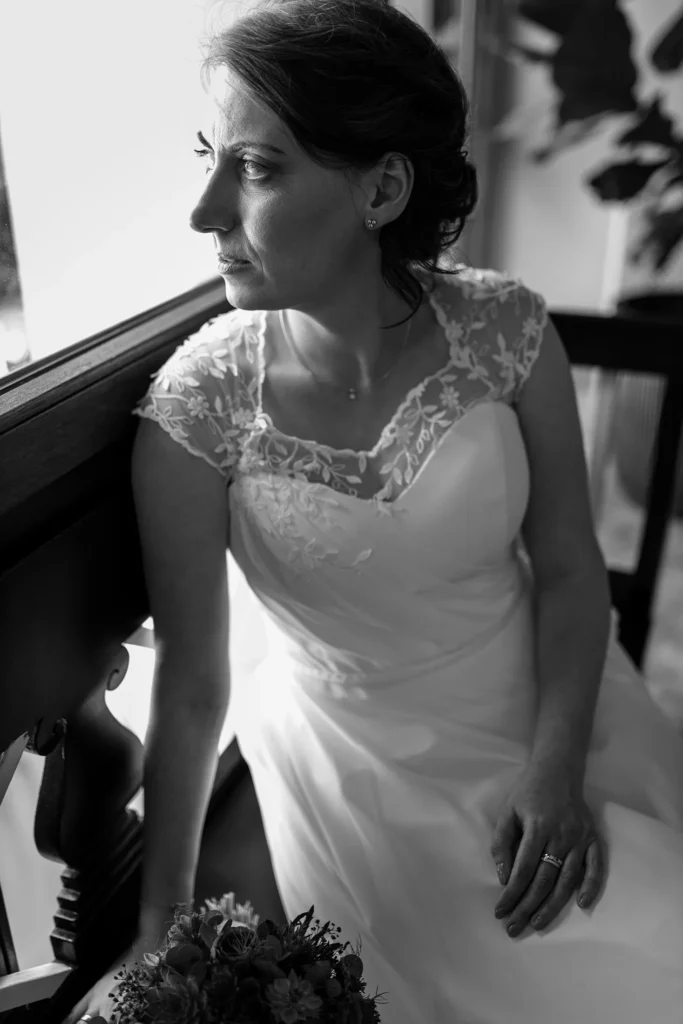 Hochzeitsfotograf Trier Luxemburg - Schwarz-weiss Aufnahme der Braut, wie sie im Hotelflur auf einer Bank sitzt, wartet und aus dem Fenster guckt.