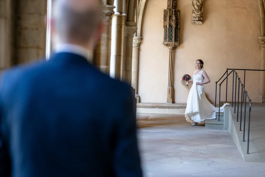 Hochzeitsfotograf Trier Luxemburg - Der First Look. Man sieht den Rücken des Bräutigams und die Braut, wie sie im Kreuzgarten des Trier Doms auf ihn zukommt.