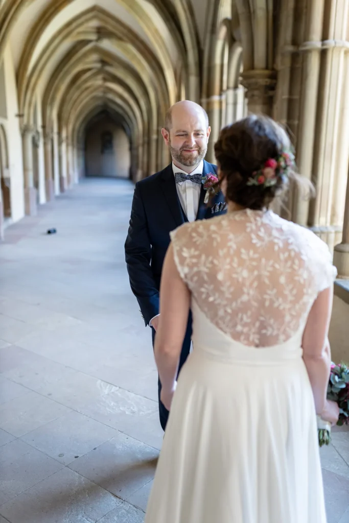 Hochzeitsfotograf Trier Luxemburg - First Look im Kreuzgarten des Trier Doms: Braut im Vordergrund von hinten, im Hintergrund sieht man die Reaktion des Bräutigams, als er seine Frau zum ersten Mal im Brautkleid sieht.
