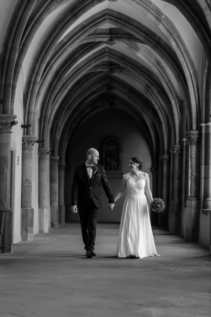 Hochzeitsfotograf Trier Luxemburg - Schwarz-weiss-Variante eines Brautpaar Bilds: Brautpaar kommt in Säulengang des Kreuzgartens im Trier Dom auf den Fotografen zu.