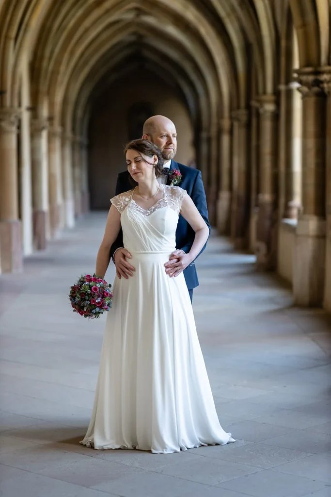 Hochzeitsfotograf Trier Luxemburg - Brautpaar-Shooting: Brätuigam steht hinter der Braut und hält sie im Arm. Beide stehen mitten in einem wunderbaren Säulengang.