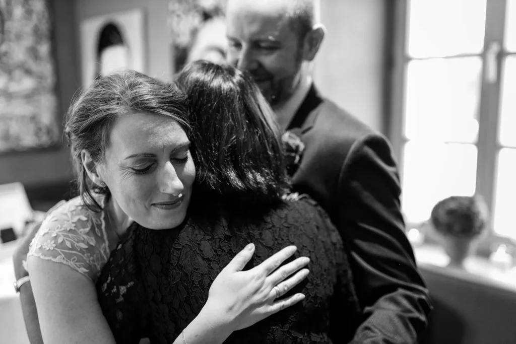 Hochzeitsfotograf Trier Luxemburg - Umarmung der Braut durch ihre Mutter. Im Hintergrund steht der Bräutigam in der Unschärfe des Fotos und guckt zu.