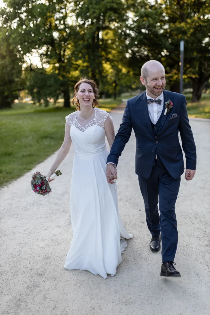 Hochzeitsfotograf Trier Luxemburg - Das lächelnde Brautpaar wird fotografiert, während es auf dem Weg des Nells Park auf den Hochzeitsfotografen zukommt, nachdem die Braut den Bräutigam mit dem Brautstrauss im Spass auf den Kopf geschlagen hat