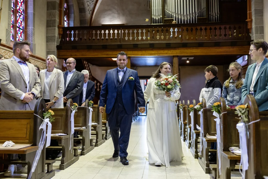 Hochzeitsfotograf Trier Luxemburg - Das Brautpaar geht beim Einzug in die Kirche durch den Mittelgang und lächelt die stehenden Gäste dabei an.