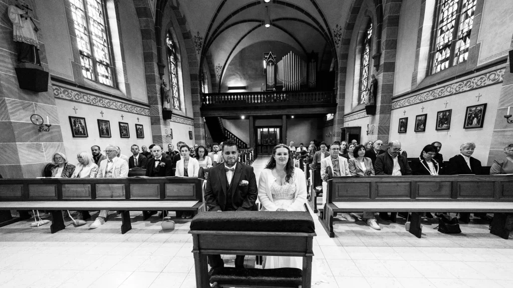 Hochzeitsfotograf Trier Luxemburg - Panorama Aufnahme schwarz-weiss: Brautpaar sitzt vorne zentral auf ihrer Bank in der Kirche. Im Hintergrund sind die Gäste auf den Bänken und das Kirchenschiff zu sehen.