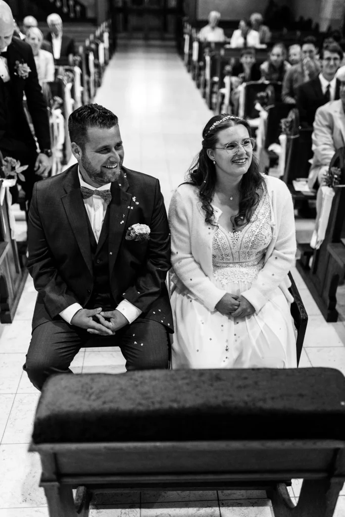 Hochzeitsfotograf Trier Luxemburg - Brautpaar sitzt zentral auf einer Bank in der Kirche. Im Hintergrund sind die Gäste auf den Bänke zu sehen. Schwarz-weiss Foto.