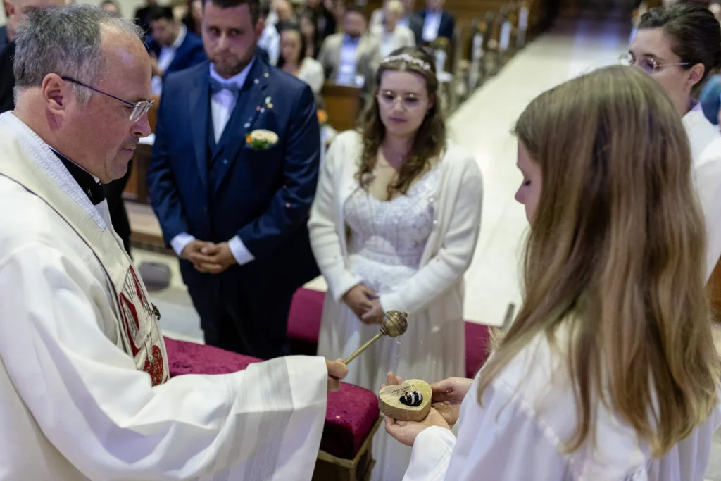 Hochzeitsfotograf Trier Luxemburg - Der Pfarrer segnt im Moment die Ringe des Brautpaars. Man sieht das Weihwasser spritzen. Im Hintergrund in der Unschärfe sieht man das Brautpaar, wie es dabei zuschaut.