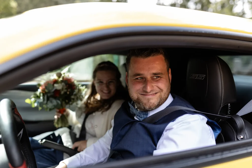 Hochzeitsfotograf Trier Luxemburg - Nahaufnahme des breit grinsenden Bräutigams, als er mit dem gelben Ford Mustang auf den Hof der Hochzeits-Location gefahren kommt.