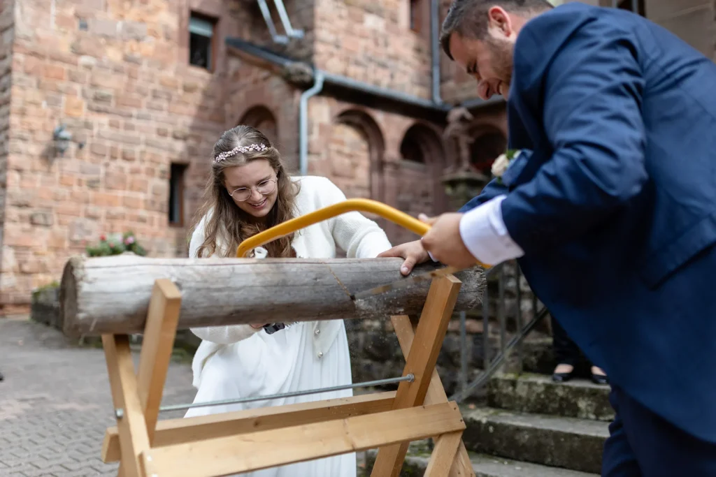 Hochzeitsfotograf Trier Luxemburg - Braut und Bräutigam beim traditionellen Baumstamm Sägen auf ihrer Hochzeit