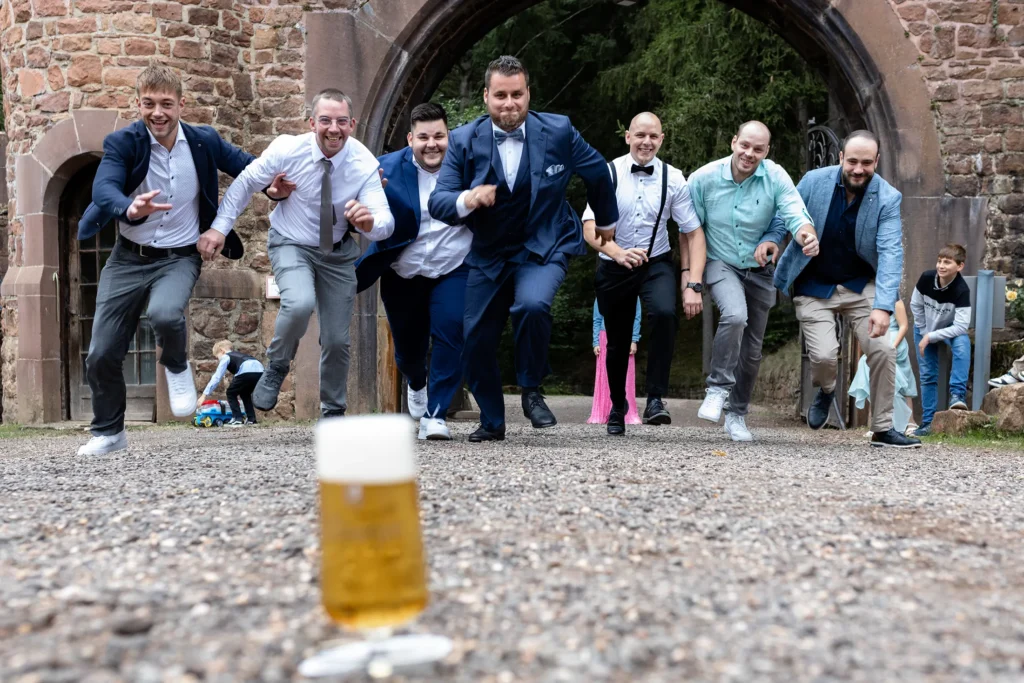 Hochzeitsfotograf Trier Luxemburg - Der Bräutigam und seine Freunde rennen auf ein volles Glas Bier zu, das auf dem Boden im Vordergrund des Fotos steht.