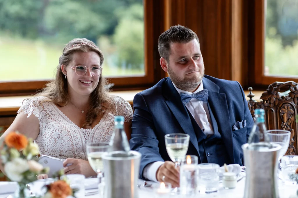 Hochzeitsfotograf Trier Luxemburg - Lächelnde Braut am Tisch, während sie einer Rede zuhört.