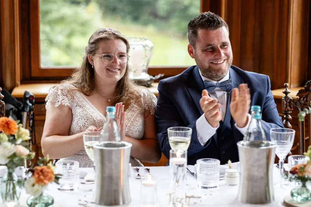 Hochzeitsfotograf Trier Luxemburg - Das Brautpaar sitzt am Tisch und klatscht Beifall zu einer Rede.