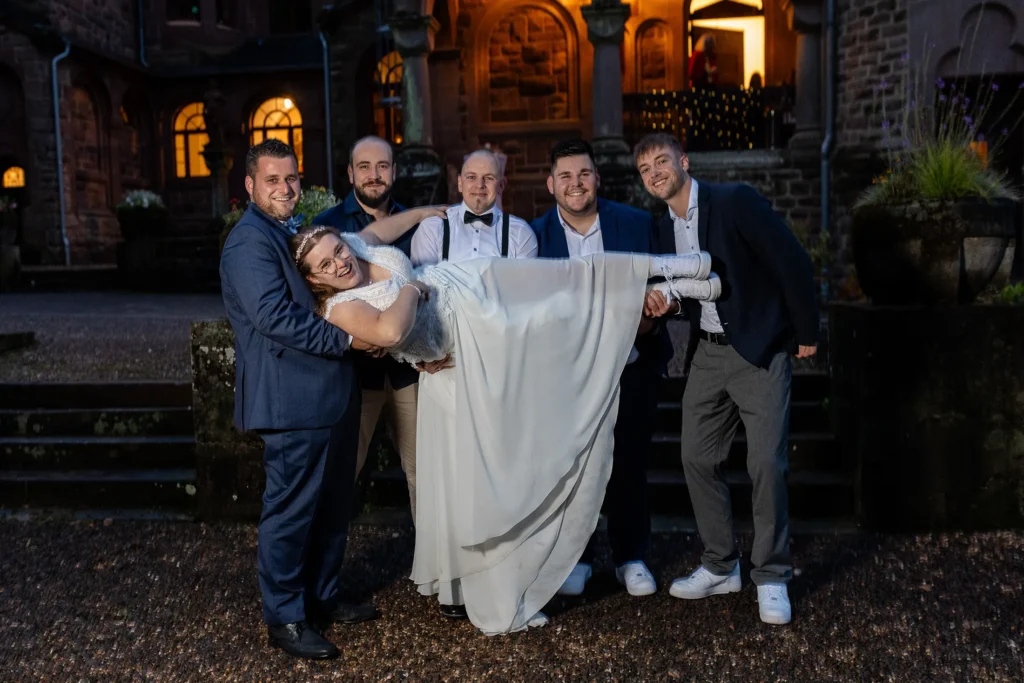 Hochzeitsfotograf Trier Luxemburg - Die Braut wird vor dem Schloss von ihrem Bräutigam und seinen Freunden getragen und lacht dabei.