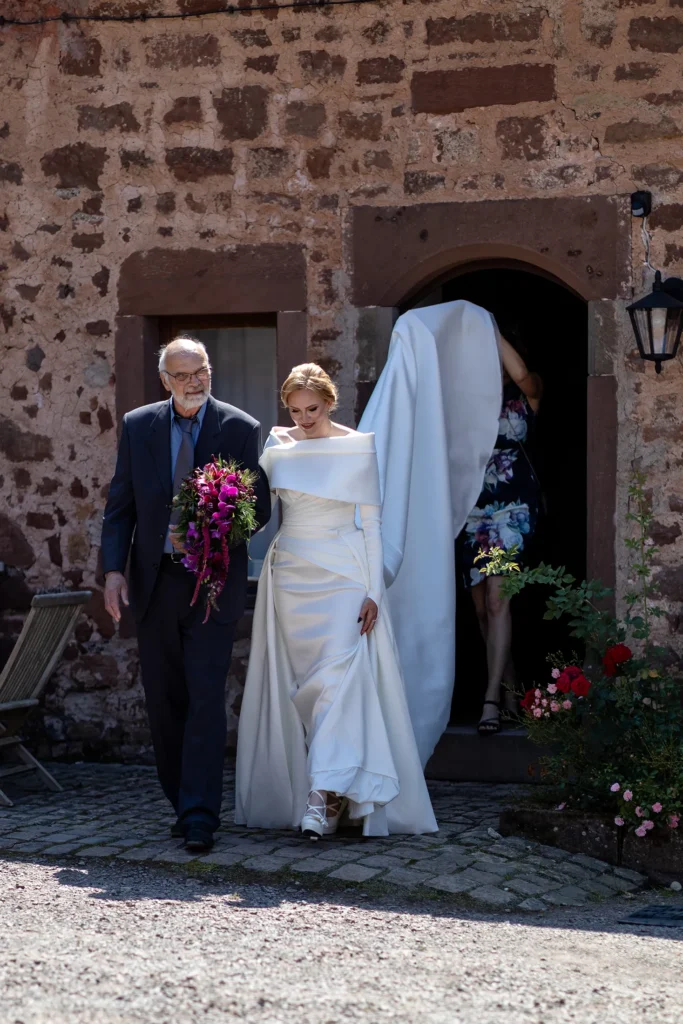 Hochzeitsfotograf Trier Luxemburg - Braut mit ihrem Vater auf dem Weg zum First Look. Trauzeugin hält das Kleid hoch