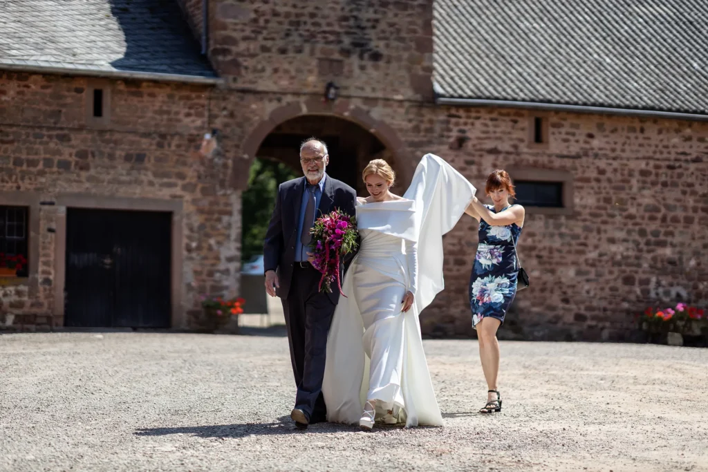 Hochzeitsfotograf Trier Luxemburg - Trauzeugin hält das Kleid hoch, während Braut und ihr Vater über den Hof von Schloss Hamm in Richtung Kapelle gehen
