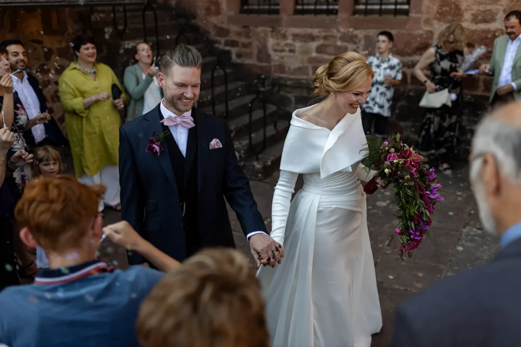 Hochzeitsfotograf Trier Luxemburg - Das lachende frisch vermählte Brautpaar kommt gerade aus der Kapelle und die Gäste um sie herum erzeugen Seifenblasen zum Empfang.