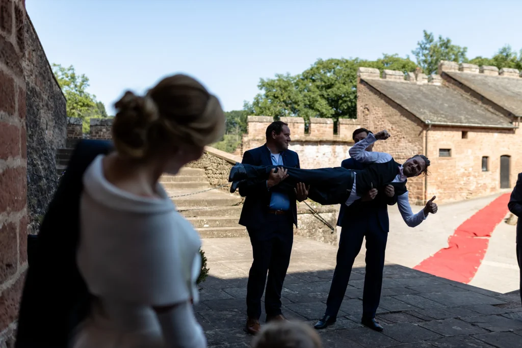 Hochzeitsfotograf Trier Luxemburg - Der Bräutigam wird von 2 Mann hochgehoben, während die Braut zusieht
