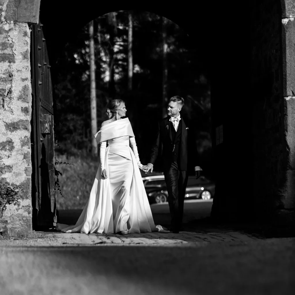 Hochzeitsfotograf Trier Luxemburg - Schwarz-weiss Aufnahme: Braut und Bräutigam halten Händchen, sehen sich an und spazieren gemeinsam durch dass Tor von Schloss Hamm